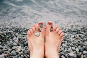 Trouver l’équilibre entre santé mentale et santé physique : la vraie clé du bien-être Close-up of female feet with stones on pebbled beach by the water, evoking relaxation and tranquility.