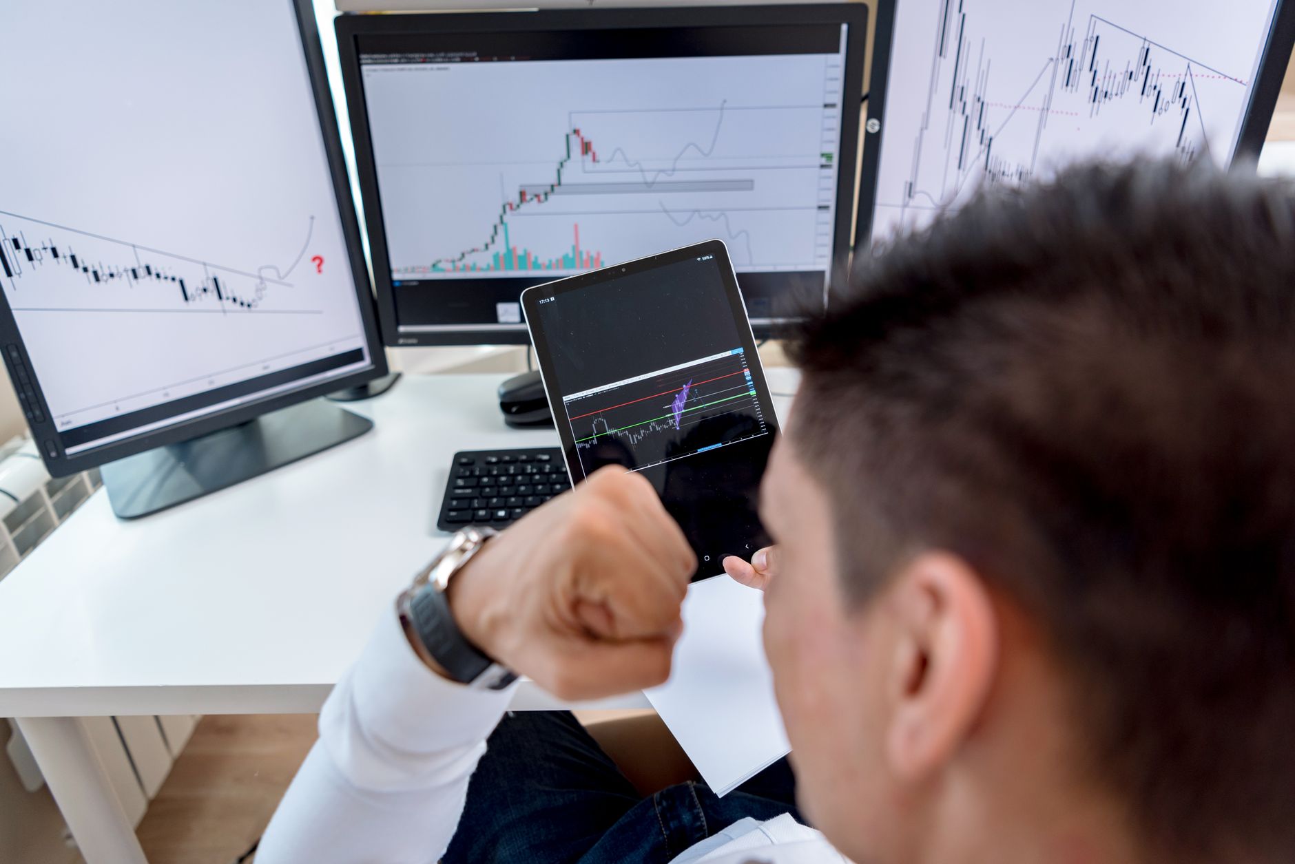 Homme vu de dos devant trois écrans et une tablette affichant des graphiques financiers en chandeliers, assis à un bureau blanc.