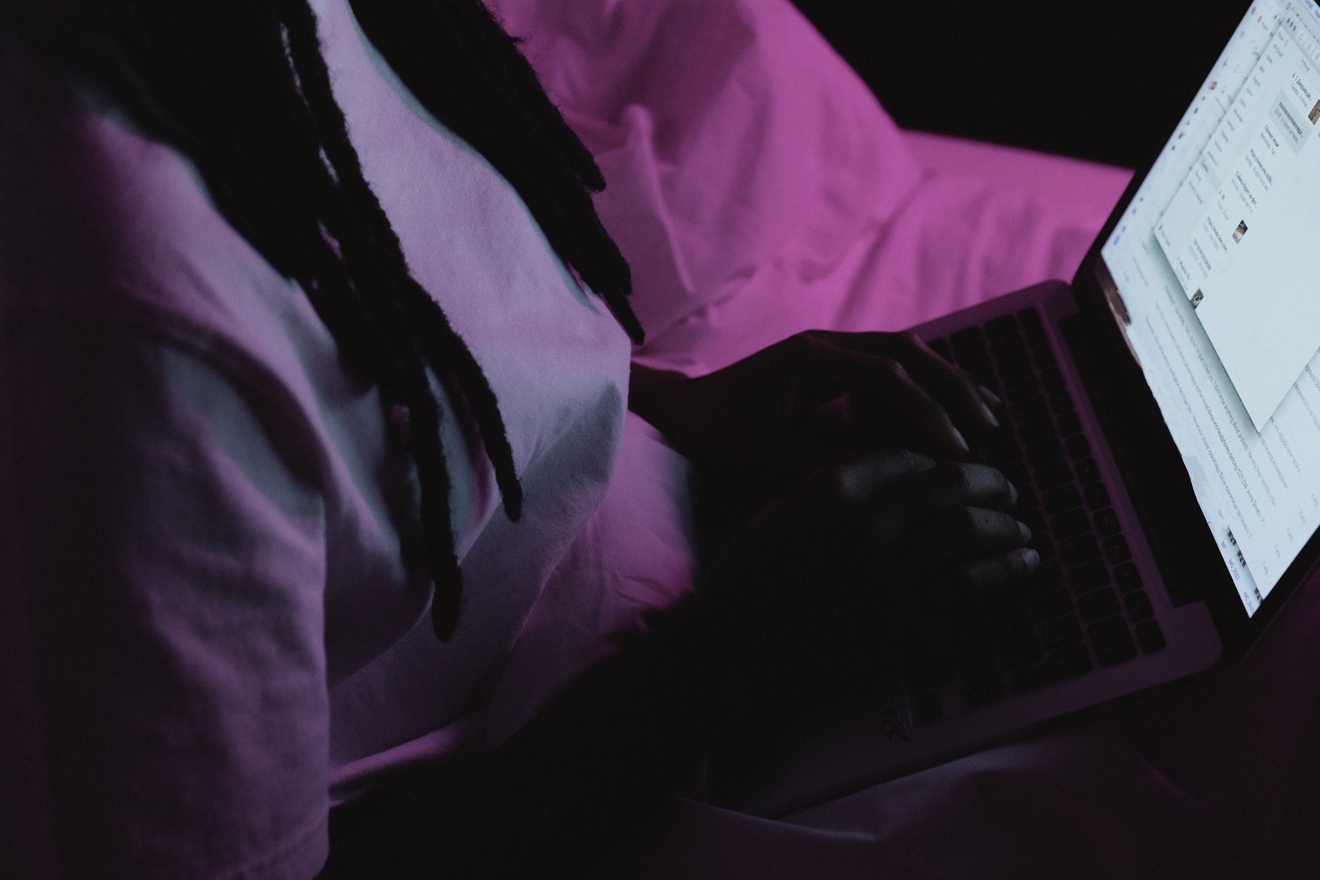 Personne aux cheveux tressés tapant sur un ordinateur portable allumé, sur un lit aux draps roses, dans une pièce sombre.