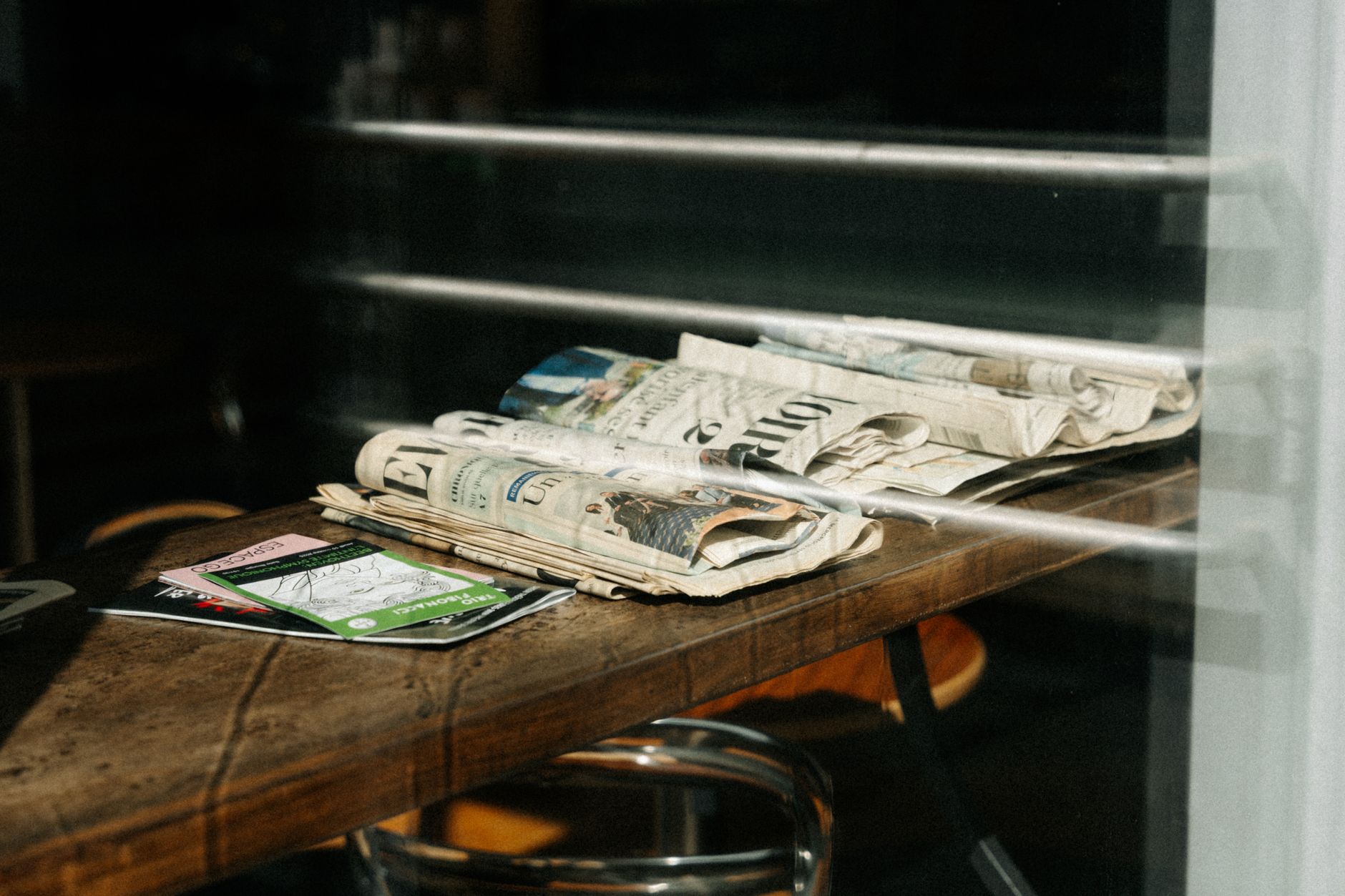 Pile de journaux imprimés posée sur une table en bois, vue à travers une vitre, avec des dépliants colorés à gauche et un tabouret en dessous.