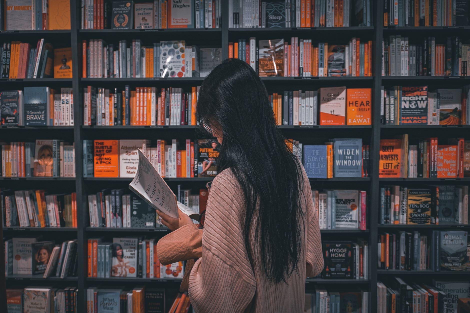 Une femme vue de dos, aux longs cheveux noirs, lit un livre ouvert devant de hauts rayonnages remplis de livres.